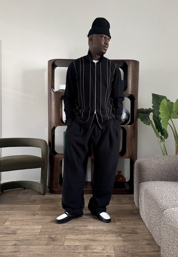 Muž oblečený v čiernom oblečení s vertikálnymi pruhmi a vysokou čiapkou stojí v interiéri pri drevenej poličke, zelenej stoličke a sivej pohovke.