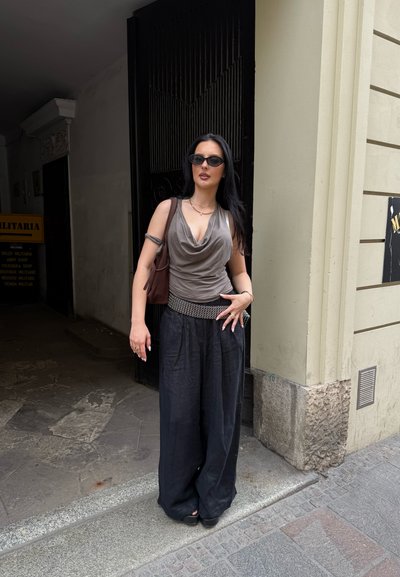 Top drapeado gris, pantalones de pierna ancha negros y un cinturón decorativo. Los accesorios incluyen gafas de sol pequeñas negras y un bolso marrón. Fondo rocoso.