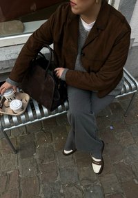 Καφέ σουέτ λευκό πάνω από γκρι πουλόβερ, γκρι παντελόνι και καφέ-λευκά παπούτσια. Μαύρη δερμάτινη τσάντα και κούπες καφέ σε δίσκο.