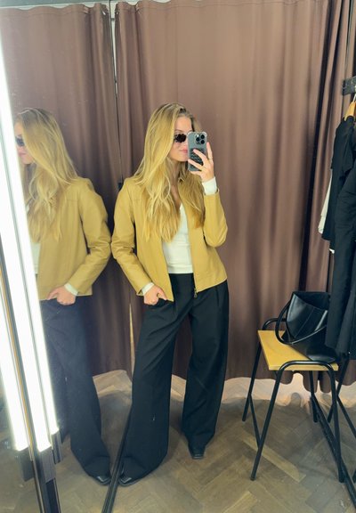 Chaqueta de cuero marrón claro junto a un espejo, sobre una blusa blanca, combinada con pantalones negros holgados. En la esquina se ven un bolso negro y una silla.