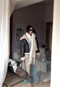 Geantă de mână din piele maro, jachetă căptușită cu blană neagră, pulover gri, blugi largi de culoare bleu deschis, eșarfă albă cu franjuri. Setare în interior.