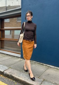 Brun genser med høy hals, oransje kordfløyelskjørt, og patentlær brune hæler. Beige skulderveske og briller. Utendørs urbant miljø.