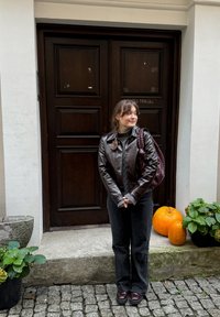 Bruine leren jas, grijze trui, donkere broek, bordeauxrode laarzen. Potplanten in de buurt, oranje pompoenen aan de voet, houten deur op de achtergrond.