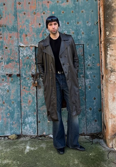 Hombre con gorro negro, gafas de sol en la cabeza, abrigo largo de cuero negro, camisa negra, vaqueros de pierna ancha, de pie frente a una puerta de metal verde envejecida.
