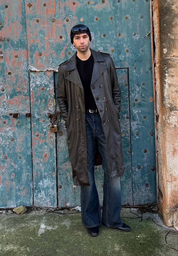 Uomo con berretto nero, occhiali da sole sulla testa, lungo cappotto nero di pelle, camicia nera, jeans a gamba larga, in piedi davanti a una porta di metallo verde usurata.