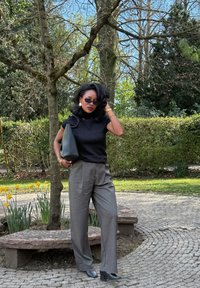 Top preto ajustado, calças largas cinza sob medida, bolsa preta, óculos de sol escuros, joias douradas. Cenário ao ar livre com vegetação e flores.