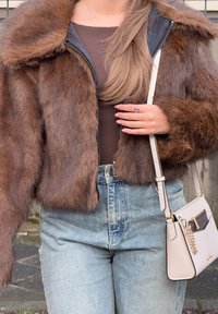 Giacca cropped in finta pelliccia marrone con una texture morbida, abbinata a jeans in denim blu a vita alta e una piccola borsa a tracolla beige.