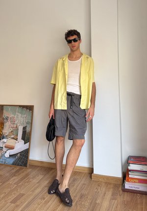 Camisa amarilla de manga corta sobre una camiseta blanca sin mangas, pantalones cortos de cargo grises, zapatos abiertos marrones, y una bolsa negra. Diseño minimalista y estilo casual.