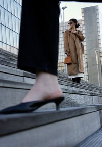 Mules nere con tacco su gradini in legno, con una figura sfocata in un cappotto marrone e una borsa arancione che sale sullo sfondo.