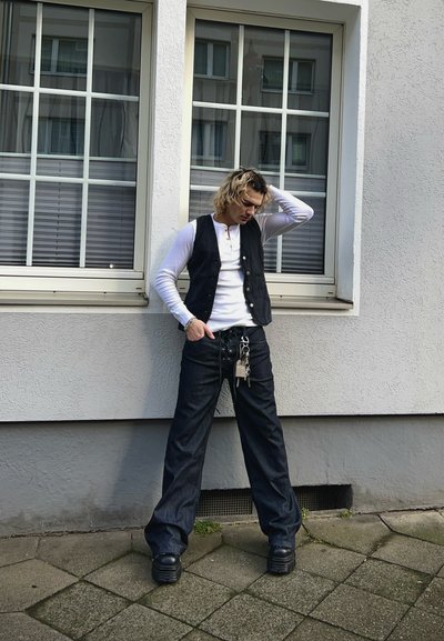 Chaleco de mezclilla negro sobre una camisa blanca de manga larga, combinado con unos vaqueros oscuros amplios con cordones en la parte delantera y zapatos de plataforma negros gruesos. Accesorios plateados.