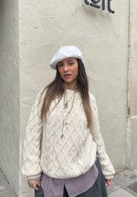 Maglione lavorato a maglia di colore crema chiaro con un motivo a rombi, abbinato a una camicia a quadri e una gonna scura. Un berretto bianco e collane a strati arricchiscono il look.