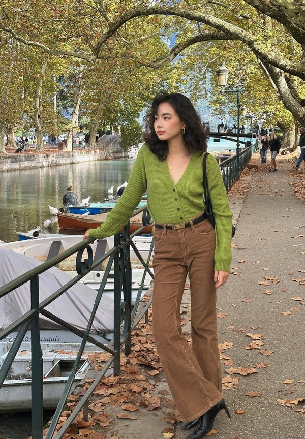 Une femme en cardigan vert et pantalon brun s’appuie sur une rambarde au bord de la rivière, près de bateaux amarrés et sous des arbres d’automne.