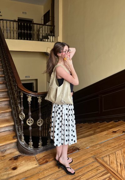 Bolso de lona en beige claro, falda de lunares en blanco y negro, con sandalias planas. Escalera y barandilla de madera en el fondo.