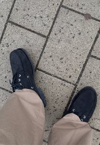 Chaussures en daim noir avec des lacets blancs et des semelles en caoutchouc, vues de dessus. Pantalon beige visible. La surface est un pavage en béton texturé.