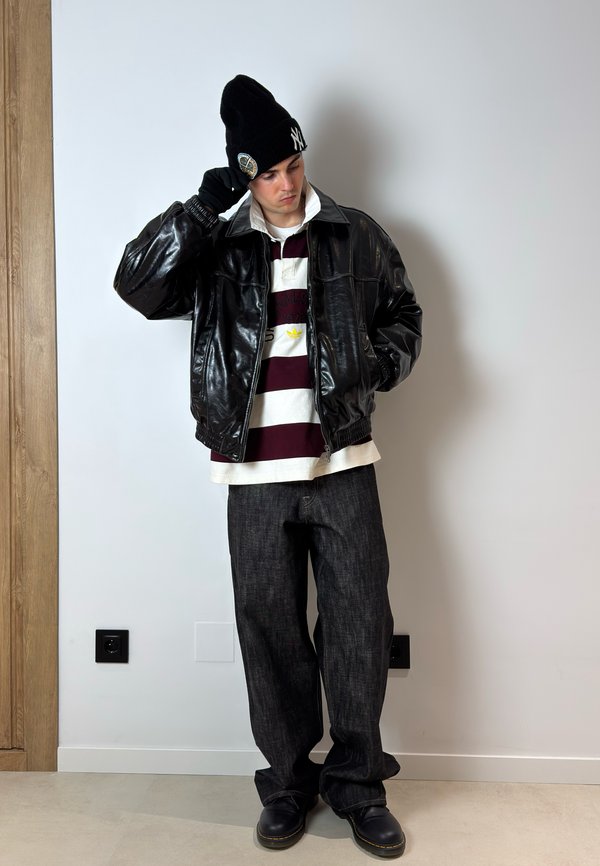 Ung mand iført sort hue, sort læderjakke, stribet poloshirt, løstsiddende mørke jeans og sorte støvler, stående mod en hvid væg.