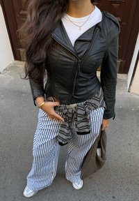 Jaqueta de couro preta com fecho de zíper, camisa branca canelada por baixo, calças largas listradas e uma gravata preta com padrão à volta da cintura. Saco castanho.