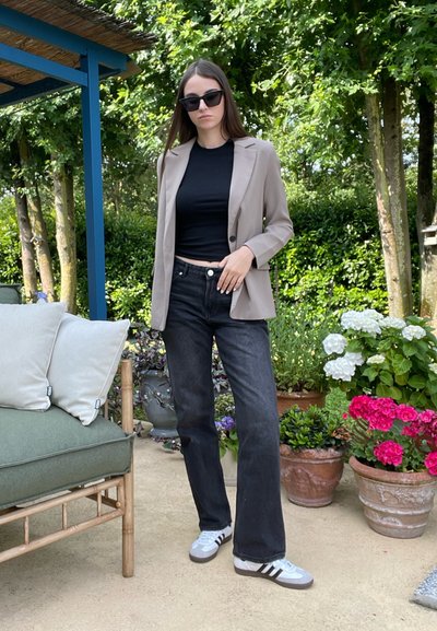 Blazer beige, top negro ajustado y jeans negros de pierna ancha. Zapatillas grises con rayas blancas. Entorno al aire libre con plantas y flores verdes.