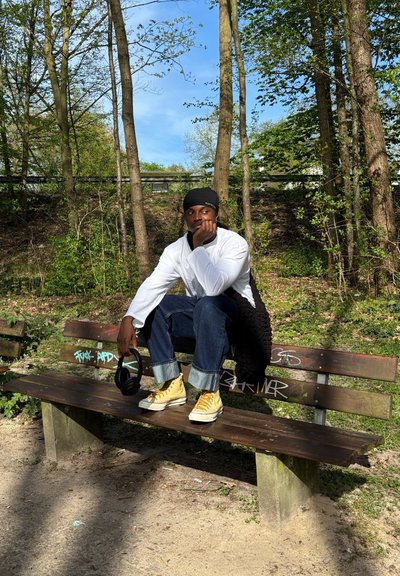 Una persona está sentada en un banco de madera, vistiendo una camisa blanca, jeans azules con dobladillos y zapatos deportivos amarillos. Sostiene unos auriculares con una mano.