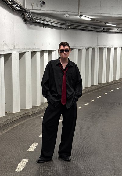 Camisa negra de gran tamaño y pantalones de pierna ancha, con una corbata burdeos y gafas de sol oscuras, de pie en un área de aparcamiento subterráneo texturizada.