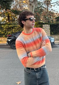 Jeune homme portant des lunettes de soleil noires et un pull coloré orange-bleu, debout les bras croisés dans la rue avec une voiture et des arbres derrière lui.