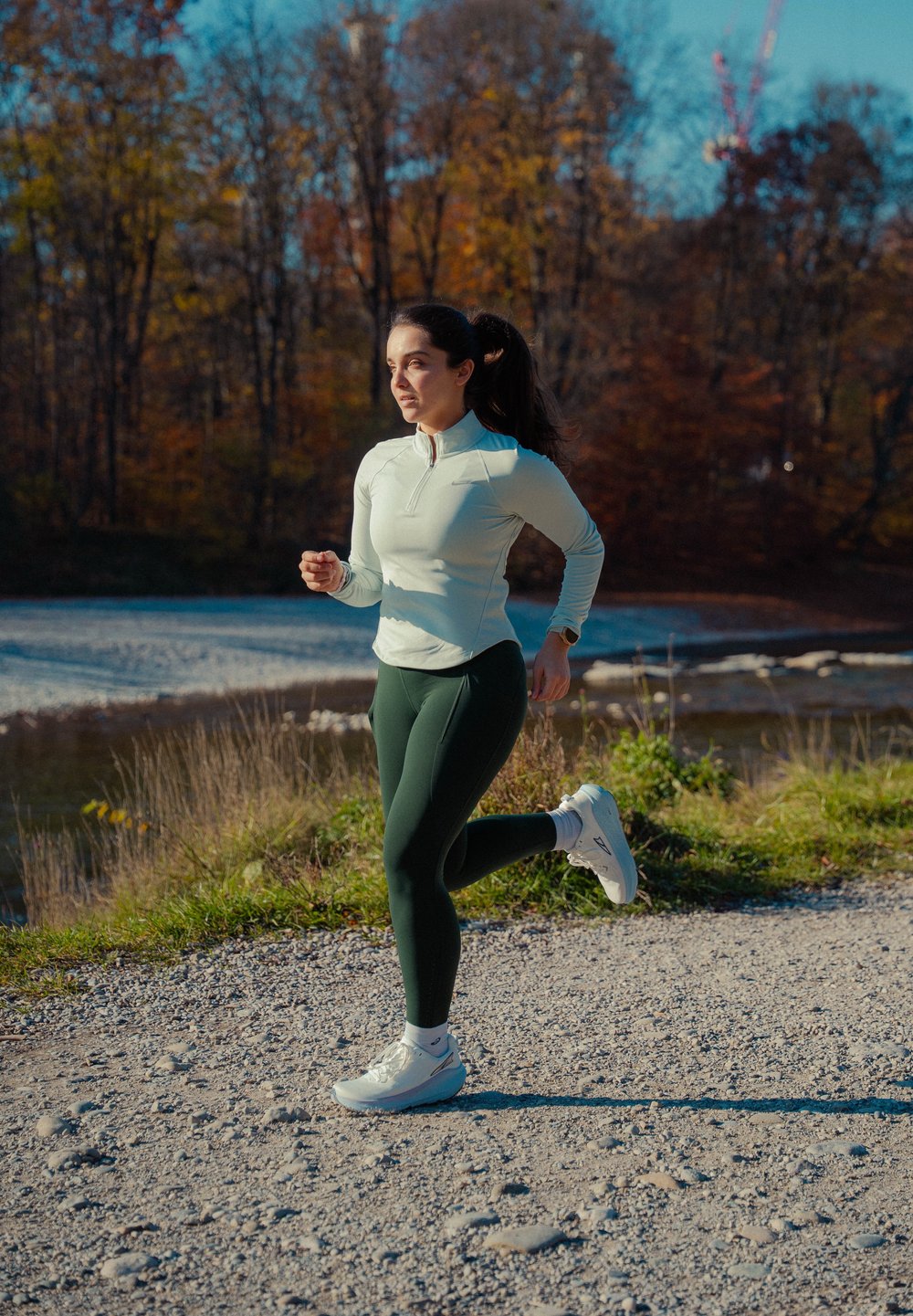 Nő kocog kavicsos ösvényen a folyó mellett, fehér hosszú ujjú felsőt, sötétzöld leggingset és fehér sportcipőt viselve, őszi környezetben.