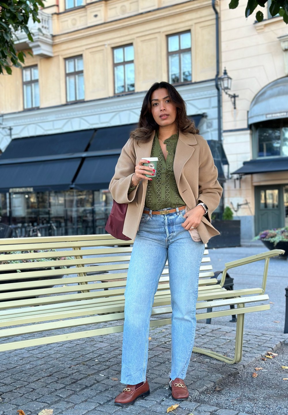 Καφέ παλτό από μαλλί, ελιά πράσινο μπλούζα με διακοσμητικές λεπτομέρειες, ανοιχτό μπλε τζιν, καφέ λουστρίνια με αλυσίδες, κρατώντας ένα φλιτζάνι καφέ.