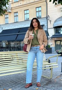 Καφέ παλτό από μαλλί, ελιά πράσινο μπλούζα με διακοσμητικές λεπτομέρειες, ανοιχτό μπλε τζιν, καφέ λουστρίνια με αλυσίδες, κρατώντας ένα φλιτζάνι καφέ.