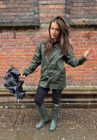 Giacca impermeabile verde con chiusura a bottoni, leggings neri e stivali di gomma verdi fino al ginocchio, che tiene un ombrello nero rovesciato contro un muro di mattoni.