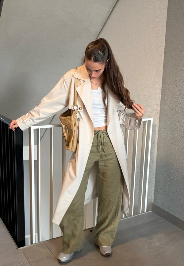 Jeune femme aux longs cheveux portant un trench beige, un crop top blanc, un pantalon large vert et des baskets, debout près de la rampe d'escalier à l'intérieur.