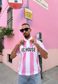 Biała koszulka polo w pionowe różowe paski z zielonym akcentem. Z przodu znajduje się czarny logo "F.C.HOUSE". Noszona z dżinsowymi szortami.