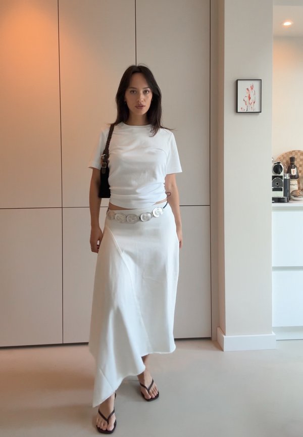 T-shirt en coton blanc associé à une jupe asymétrique blanche. Accentué par une ceinture en argent et un sac à épaule noir. Porté avec des tongs noires.