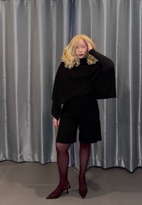 Cape noire portée sur un haut noir, associée à un short noir. Collants transparents bordeaux et talons brillants bordeaux. En arrière-plan, des rideaux gris.