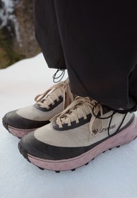 Personne portant des baskets beige et noires avec des semelles roses, debout sur la neige à l'extérieur, vêtue d'un pantalon noir.