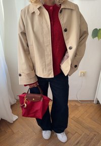 Pessoa a usar um casaco bege, suéter vermelho, calças pretas e ténis brancos, segurando uma bolsa vermelha com um chaveiro em forma de cereja.