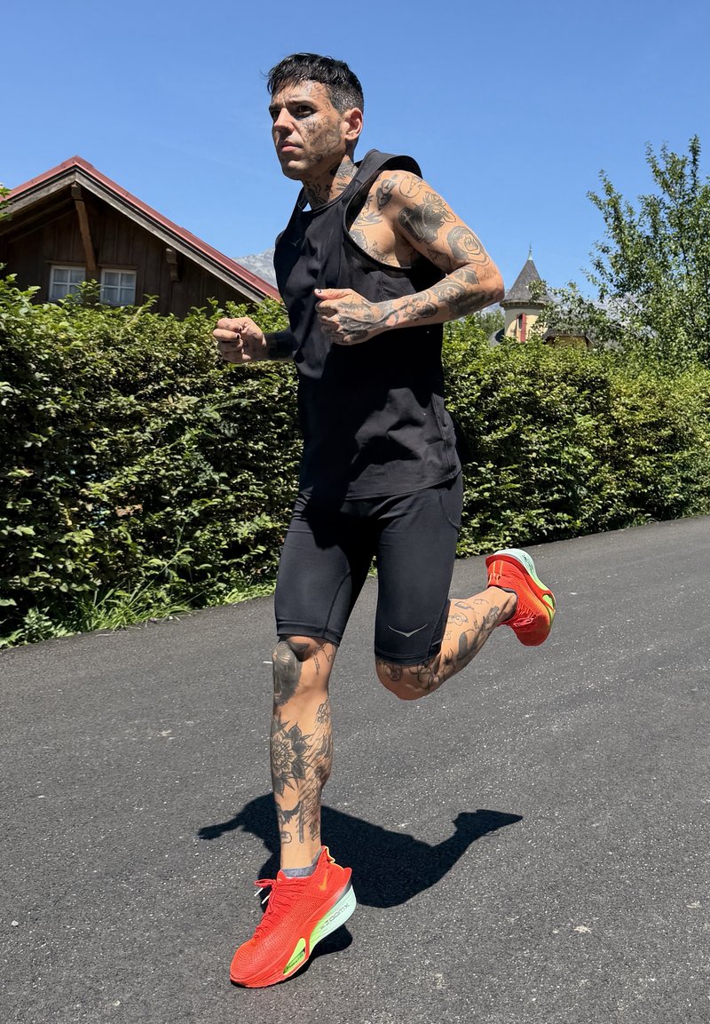 Tatuażysta w czarnych ubraniach sportowych i jaskrawo pomarańczowych butach do biegania jogginguje na zewnątrz po utwardzonej drodze obok zieleni i domów.