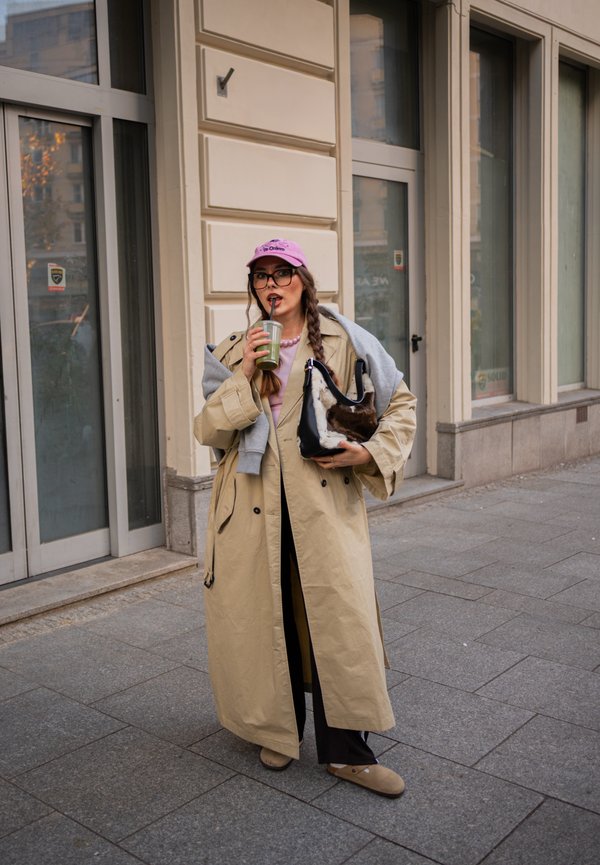 Una mujer con una gabardina beige y una gorra rosa bebe con una pajita mientras sostiene a un perro pequeño frente a una tienda.