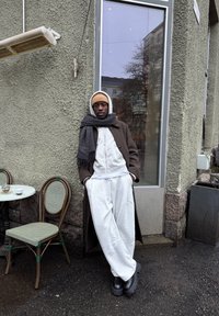 Sweat-shirt blanc à capuche, pantalon de jogging gris, manteau long marron foncé, bottes à plateforme noires. Accessoires incluent une écharpe grise et un bonnet beige.