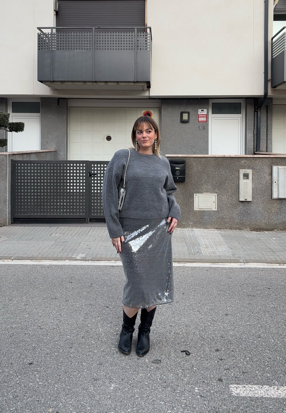 Γυναίκα που στέκεται σε δρόμο φορώντας γκρι πουλόβερ, ασημένια midi φούστα με παγιέτες, μαύρες μπότες και ασημένια τσάντα με αλυσίδα, με κατοικημένα κτίρια στο φόντο.