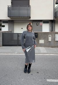 Γυναίκα που στέκεται σε δρόμο φορώντας γκρι πουλόβερ, ασημένια midi φούστα με παγιέτες, μαύρες μπότες και ασημένια τσάντα με αλυσίδα, με κατοικημένα κτίρια στο φόντο.