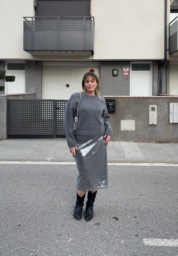 Frau steht auf der Straße, trägt einen grauen Pullover, einen silbernen Pailletten-Midirock, schwarze Stiefel und eine silberne Kettentasche, mit Wohngebäuden im Hintergrund.