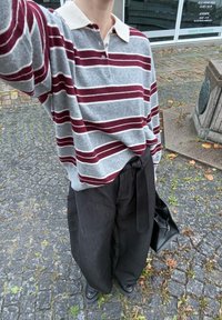 Szara koszula z długim rękawem w bordowe paski oraz kremowym kołnierzykiem, w połączeniu z luźnymi czarnymi spodniami i czarną torbą. Teksturowane tło z kostki brukowej.