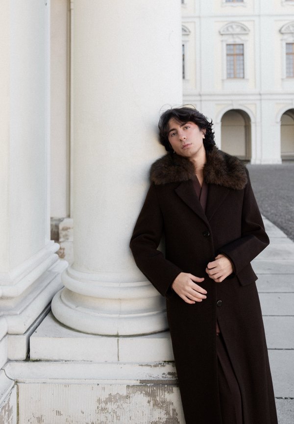 Persona con cappotto marrone scuro con collo di pelliccia appoggiata a una grande colonna bianca all'esterno di un edificio classico con archi e finestre.