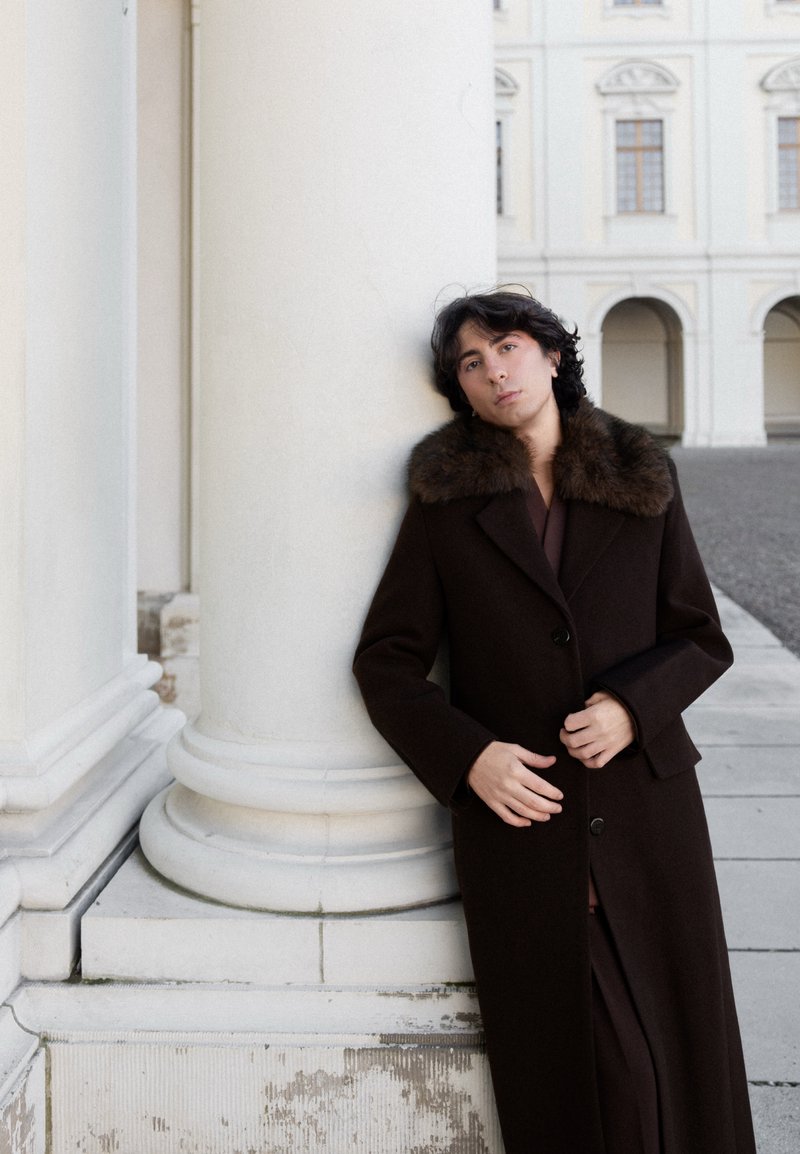 Personne en manteau brun foncé avec col en fourrure appuyée contre une grande colonne blanche à l'extérieur d'un bâtiment classique avec des arches et des fenêtres.