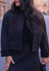 Veste noire courte avec fermeture à bouton, texture douce et manches retroussées, associée à une jupe ajustée assortie. Détail flou subtil sur le dessus.