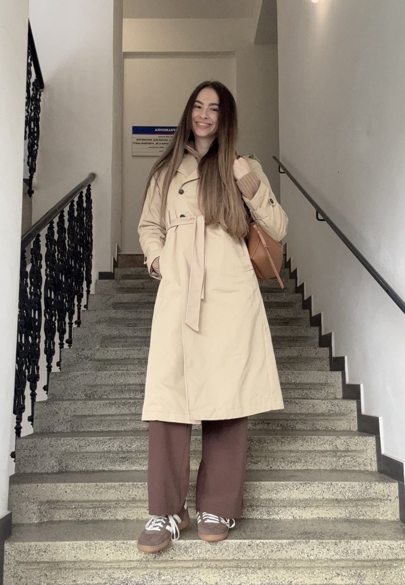 Žena s dlhými hnedými vlasmi v béžovom trenčkotovom kabáte, hnedých nohaviciach a teniskách stojí na vnútorných kamenných schodoch a drží hnedú tašku cez rameno.