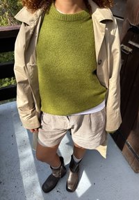 Beige trenchcoat over en grønn strukturert genser, stripete shorts og brune ankelstøvler. Støvlene har lav hæl og oppbrettet design.
