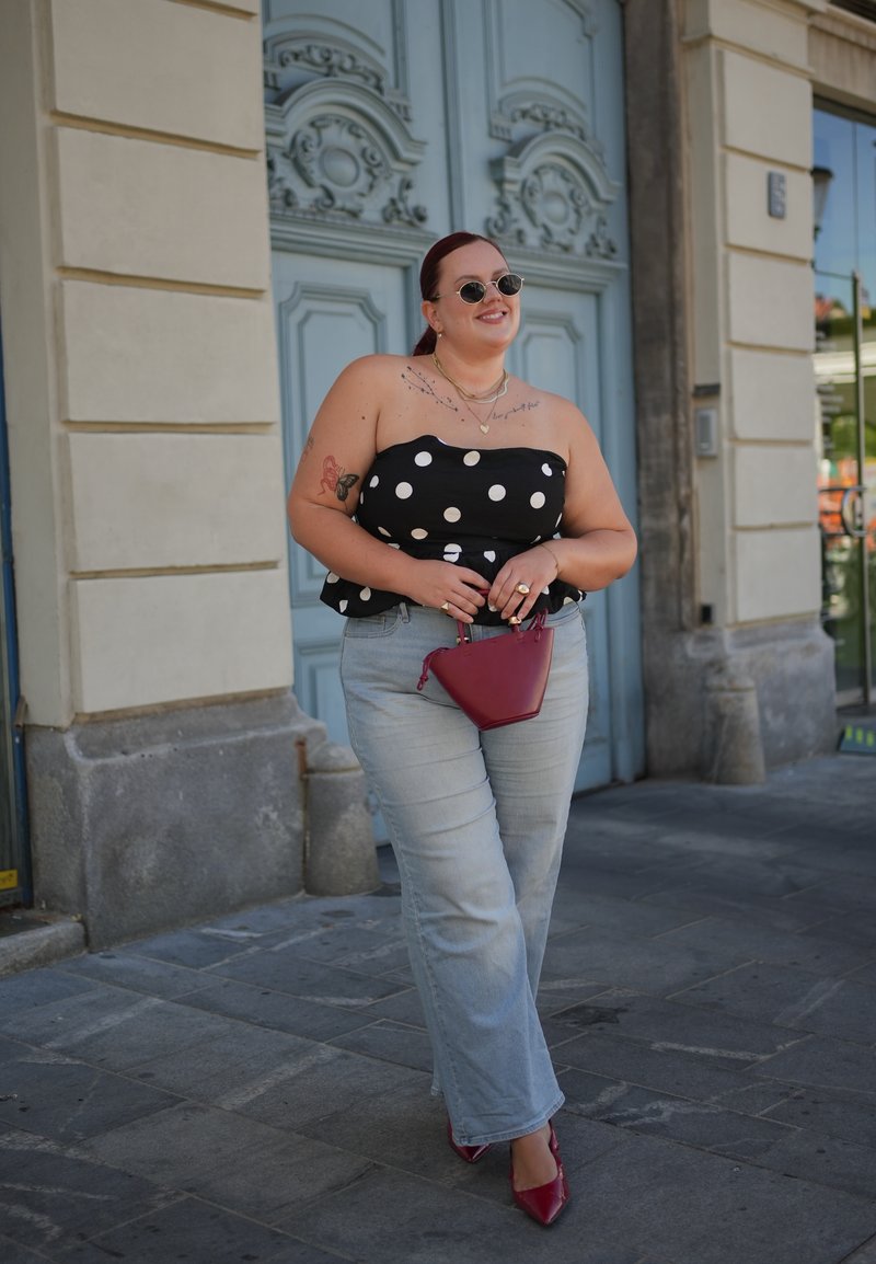 Žena so slnečnými okuliarmi, čiernym topom s bodkami, svetlými džínsami a červenými topánkami, drží malú červenú kabelku, stojí na kamennej ulici pred modrými dverami.