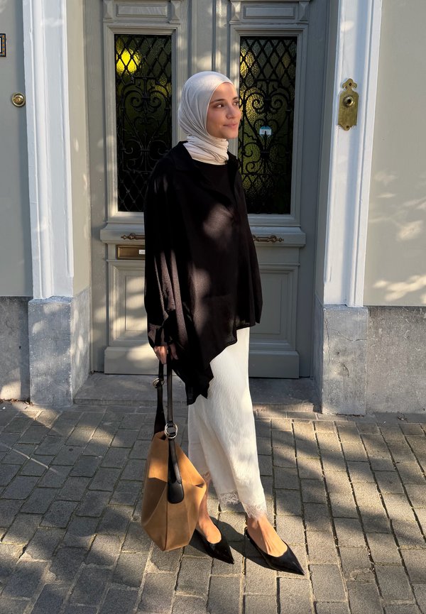 Mujer con hijab claro, camiseta negra, falda blanca, sosteniendo un bolso marrón, de pie frente a una puerta gris con paneles de vidrio decorativos.