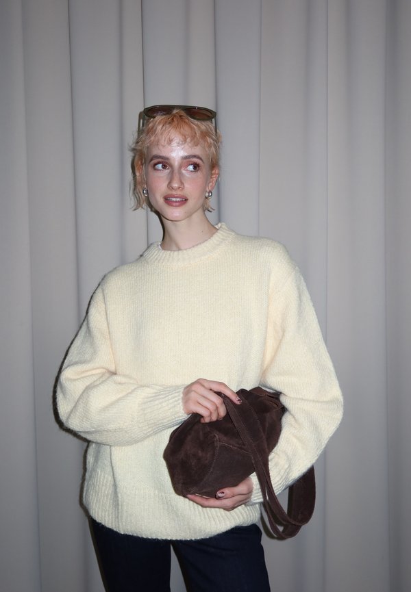 Personne aux cheveux blonds courts portant un pull crème, tenant un sac à main en daim marron, debout devant un rideau gris uni.