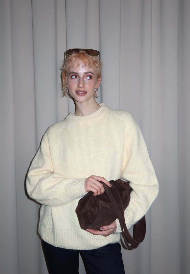 Personne aux cheveux blonds courts portant un pull crème, tenant un sac à main en daim marron, debout devant un rideau gris uni.